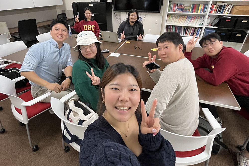 A group of international students at USC gathers around a table in a welcoming classroom setting, engaging in conversation and making peace signs. The students are participating in an ALI Conversation Group session, practicing English and discussing various topics in an informal and friendly environment.