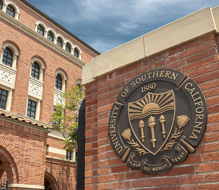 A bronze seal of the University of Southern California (USC) mounted on a red brick pillar at the entrance of the campus. The seal features three torches, a setting sun, and the Latin motto 'Palmam Qui Meruit Ferat.' In the background, a brick academic building with arched walkways and decorative white accents is visible under a bright blue sky.