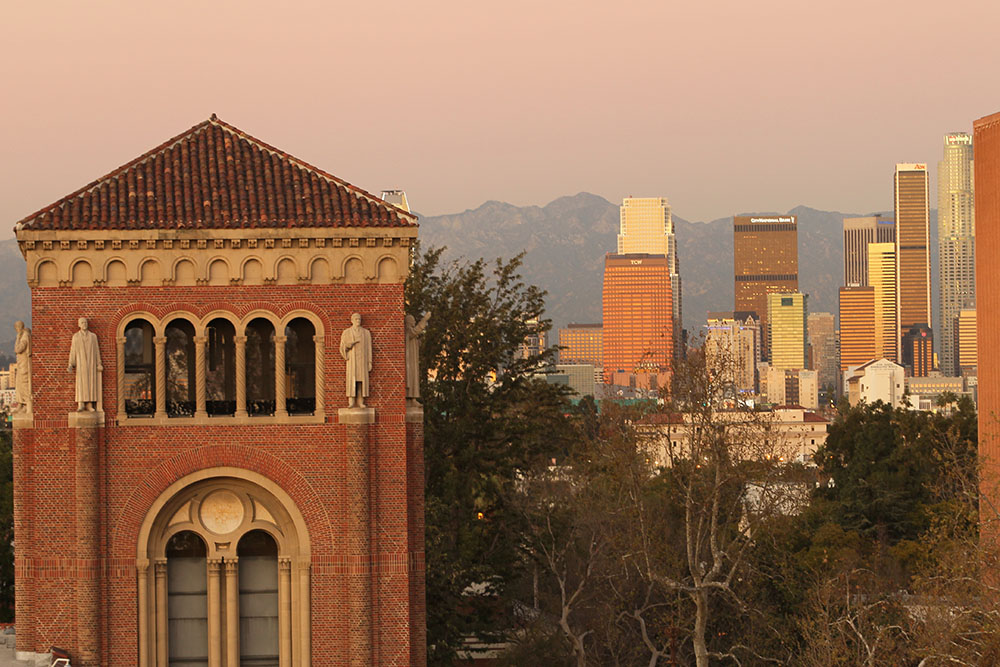 A historic red brick building at USC with arched windows and statues on its facade, set against the backdrop of the Los Angeles skyline and mountains at sunset.