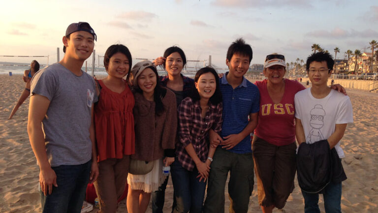 USC International Teaching Assistants (ITAs) with Program Director Lucienne enjoying a day at the beach, smiling together with ocean waves and volleyball nets in the background at sunset.