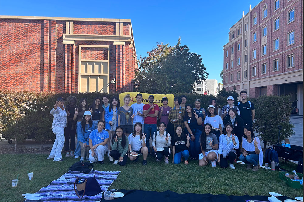 A group of USC international teaching assistants (ITAs) and undergraduate student consultants (uSCs) gathered outdoors for a picnic, posing together on a sunny day with a USC banner in the background.