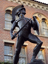 Statue of Tommy Trojan, the iconic bronze warrior figure at USC, standing with a sword and shield in front of a historic brick building.